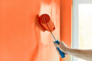 Close-up of an orange wall and a hand holding a paint roller, rolling paint on the wall.