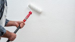 Caucasian house painter worker in white overalls, painting the wall with white paint using the roller. 